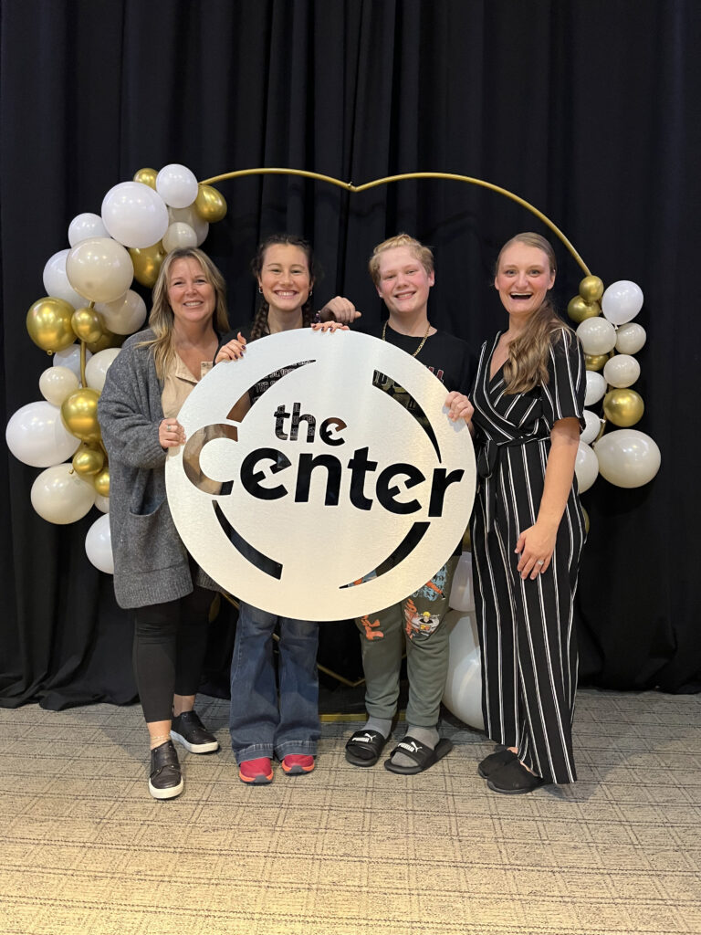 Two staff members and two students holding a large The Center sign in front of a balloon arch
