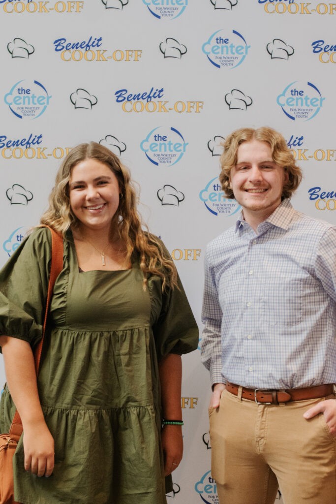 Two young adults smiling in front of The Center Benefit Cook-Off backdrop