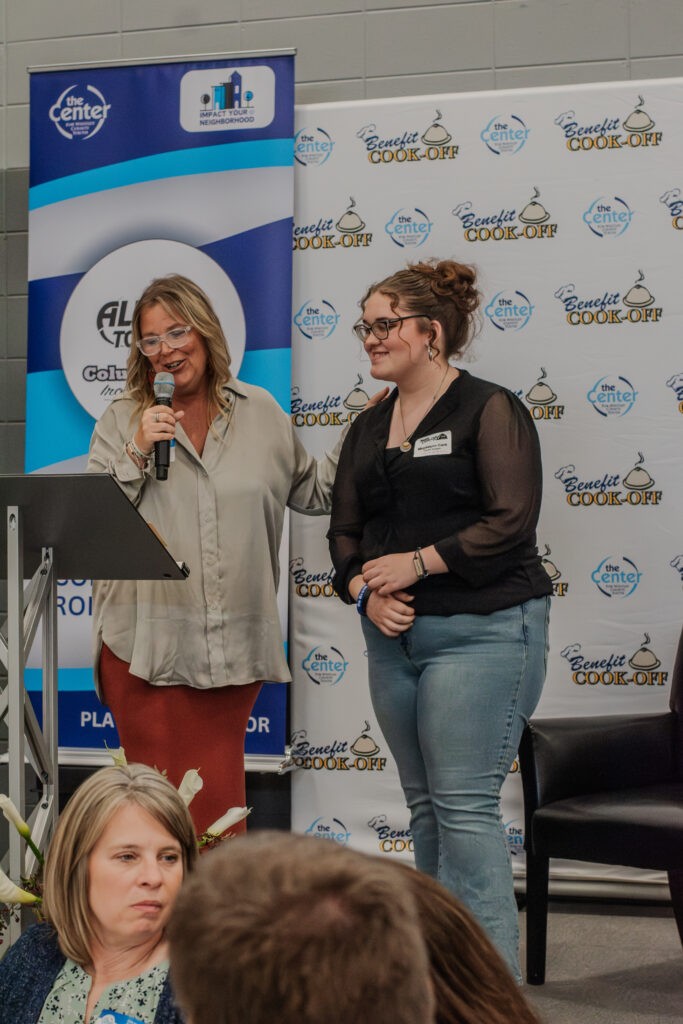 A student standing with a speaker at the microphone in front of a Benefit Cook-Off backdrop